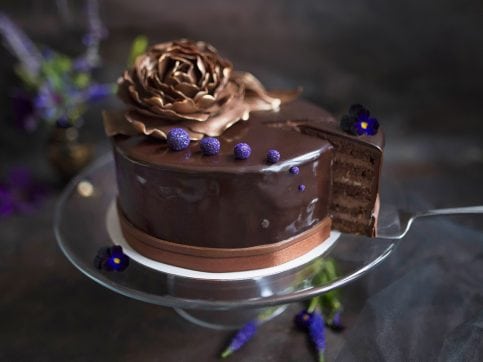Obrazek przedstawia elegancki, oblany czekoladą tort marcello udekorowany słotym kwiatem z masy cukrowej. Z tortu wykrojony jest jeden kawałek, na którym widać przekrój tortu, czyli cienkie liski biszkoptu i czekoladową masę.