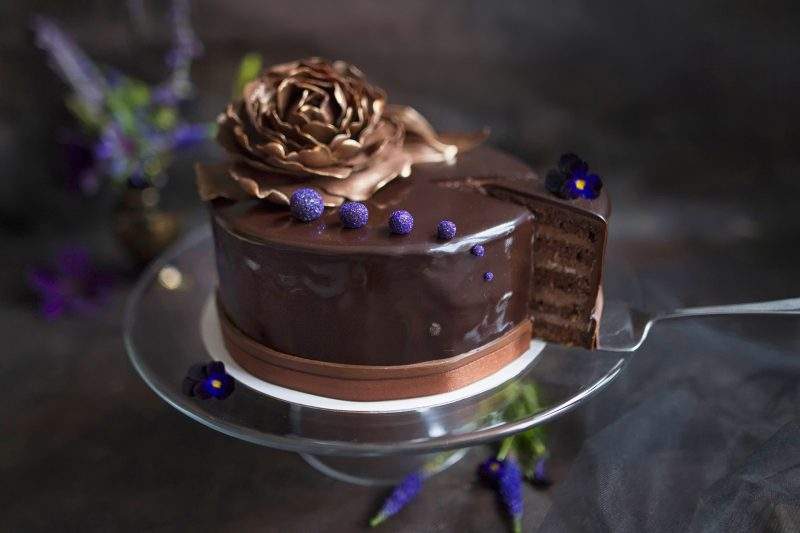 Obrazek przedstawia elegancki, oblany czekoladą tort marcello udekorowany słotym kwiatem z masy cukrowej. Z tortu wykrojony jest jeden kawałek, na którym widać przekrój tortu, czyli cienkie liski biszkoptu i czekoladową masę.
