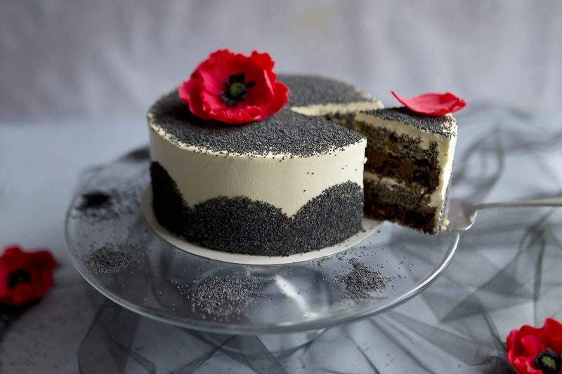 Na zdjęciu widać efektowny tort makowy. na szklanej paterze. Tort udekorowany jest ziarnami maku oraz kwiatami maku wykonanymi z cukru.